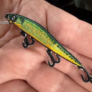 May include: Embroidered fishing lure patch featuring green, yellow, and black colors. The patch has a detailed design with three black hooks and a silver-colored lip. The patch is designed to be sewn onto clothing or accessories.