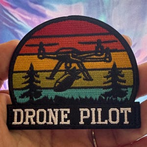 May include: Embroidered patch featuring a drone silhouette with a bomb, set against a sunset gradient of red, orange, yellow, and green. The text "DRONE PILOT" is displayed in white on a black rectangular base.