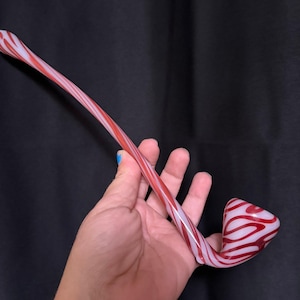 Op de afbeelding: Een handgeblazen glazen pijp met een rood-wit wervelontwerp. De pijp heeft een gebogen steel en een afgeronde kom. De kleuren zijn levendig en het ontwerp is opvallend, waardoor het een uniek stuk is.