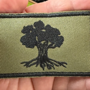 May include: Olive green rectangular patch with a black tree silhouette. The tree has a detailed trunk and roots, and a full canopy. The patch has a black border and is made of a textured fabric.