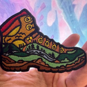 May include: Embroidered patch in the shape of a hiking boot, featuring a landscape design. The patch showcases a sunset, mountains, trees, and a river, with colors including brown, green, yellow, and orange. The boot has a black border.