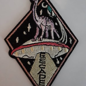 May include: Embroidered patch featuring a pink dinosaur on a UFO, set against a black diamond shape with a pink border. The UFO has a red and white design, and the patch includes text in a stylized font.