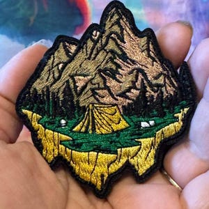 May include: Embroidered patch featuring a mountain landscape with a tent, trees, and a grassy area. The patch has a gold, green, and black color scheme, with a textured appearance. The patch is in the shape of a mountain range.