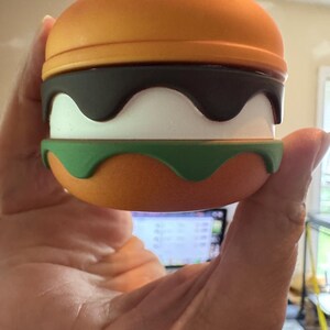 May include: A novelty burger-shaped container with an orange top and bottom, a dark brown wavy layer, a white layer, and a green base. The top has small white details. The container is held in a hand.