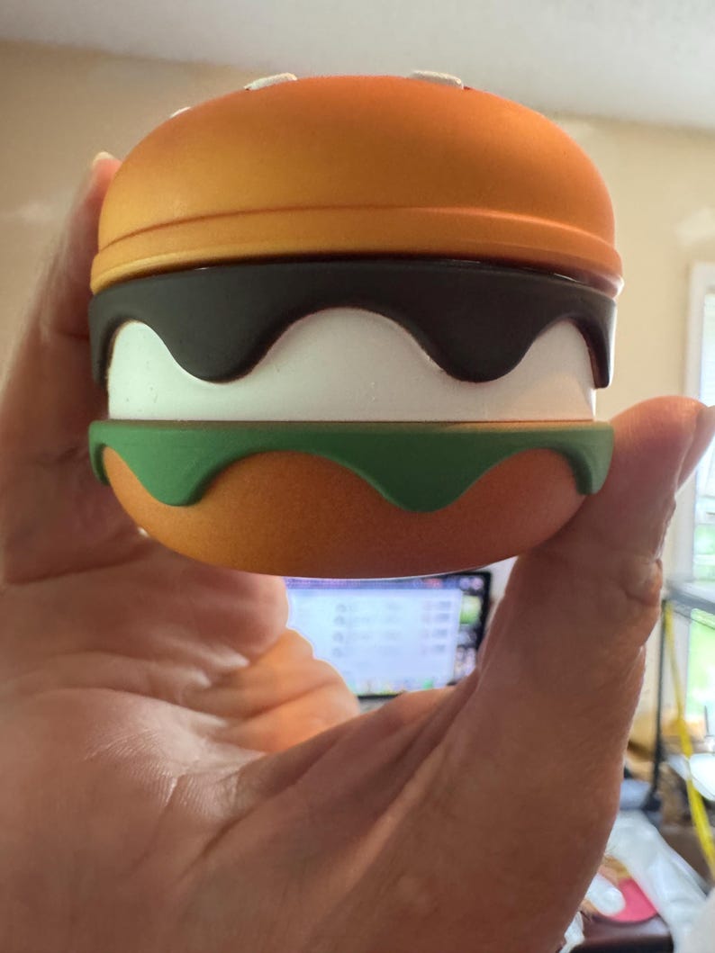 May include: A novelty burger-shaped container with an orange top and bottom, a dark brown wavy layer, a white layer, and a green base. The top has small white details. The container is held in a hand.