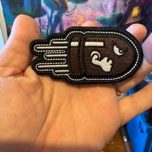 May include: A brown and black embroidered patch in the shape of a bullet with a face, white accents, and a black border. The patch has white lines to indicate motion.