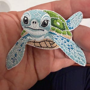 May include: Embroidered patch of a sea turtle with a light blue head and flippers, a green shell, and a tan underside. The turtle has large, expressive eyes and is held in a person's hand. The patch is likely for clothing or accessories.