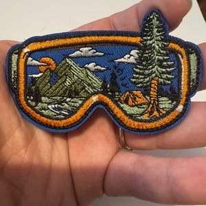 May include: Embroidered patch in the shape of goggles, featuring a scenic outdoor landscape. The patch has a blue background with a mountain range, sun, clouds, and a tent. The goggles are outlined in orange and blue.