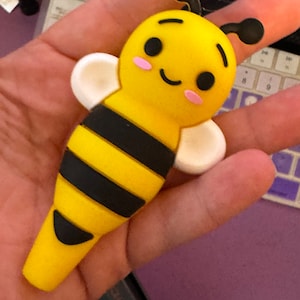 May include: A yellow and black bee-shaped decorative item with a smiling face, pink cheeks, and white wings. The bee has black stripes and antennae. The item is held in a hand.