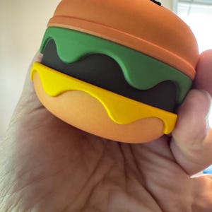 May include: A novelty burger-shaped object with orange, green, yellow, and brown layers. The top and bottom buns are orange, with a green layer resembling lettuce, a brown patty, and yellow sauce.