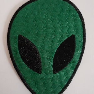 May include: Green alien head patch with black eyes and a black border. The patch is embroidered with a textured appearance. The alien head is oval-shaped and has a simple, minimalist design.