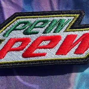 May include: Embroidered patch with the text "PEW PEW" in green and red, resembling a popular beverage logo. The patch has a black border with yellow and white accents, set against a colorful, textured background.