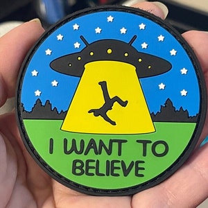 May include: A green, black, and yellow embroidered patch with a cartoon UFO and a stick figure falling from the sky. The patch says "I WANT TO BELIEVE" in black text.