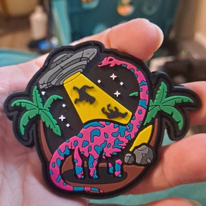 May include: A pink and blue dinosaur with a long neck stands on a brown surface with palm trees and a flying saucer. The dinosaur is surrounded by white stars.