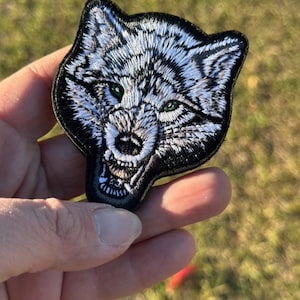May include: Embroidered patch featuring a detailed wolf head with white and black fur, green eyes, and an open mouth. The patch has a black border and is held against a blurred green background.