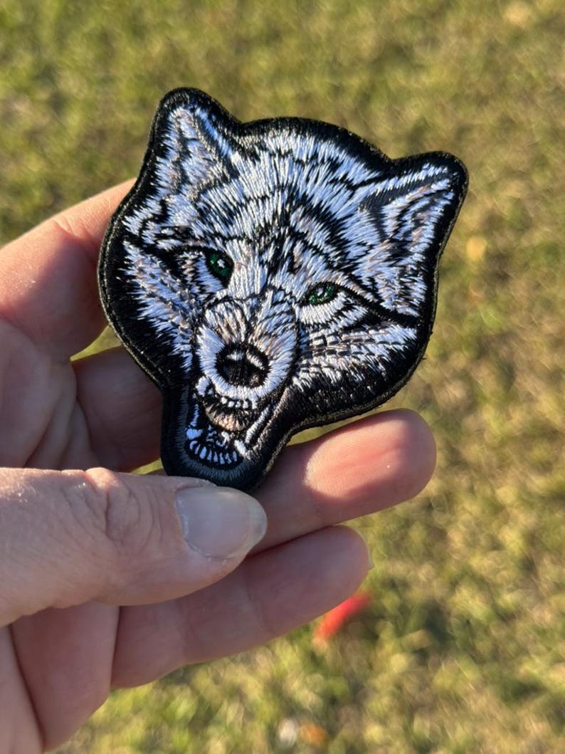 May include: Embroidered patch depicting a wolf's head with white and black fur, green eyes, and an open mouth. The patch has a black border and is held against a blurred green background.