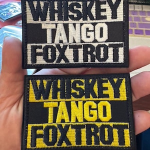 May include: Two embroidered patches with the text "WHISKEY TANGO FOXTROT". One patch has white lettering on a black background. The other has yellow lettering on a black background.
