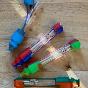 Könnte beinhalten: Vier farbenfrohe Glaspfeifen mit Silikonakzenten. Die Pfeifen haben ein klares Glasrohr mit einem Metallfilter und sind mit blauem, rotem, grünem und orangefarbenem Silikon akzentuiert. Die Pfeifen sind zum Rauchen bestimmt und auf einer Holzoberfläche angeordnet.