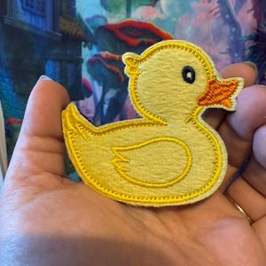 May include: A yellow duck patch with black eyes and an orange beak. The patch is outlined in yellow stitching. The duck patch is a cute, whimsical design.