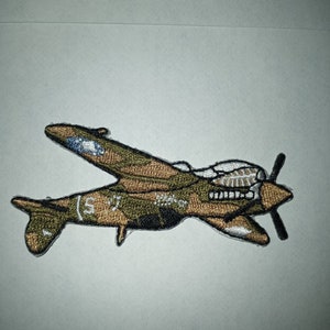 May include: A brown and green embroidered patch of a World War II era P-40 Warhawk fighter plane. The plane has a white star on the tail and the number "57" on the side. The patch is approximately 3 inches long.