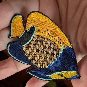 May include: Embroidered patch featuring a colorful fish design. The patch showcases a fish with a yellow body, a navy blue head and fins, and a detailed, textured pattern. The patch is likely for decorative purposes, such as on clothing or bags.