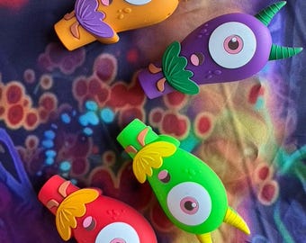May include: Four colorful, cartoonish monster-shaped objects. Each has a large, round eye with a pink iris, and a small, pointed horn. The objects are in orange, purple, green, and red, with matching accent colors. The objects are likely toys or decorative items.