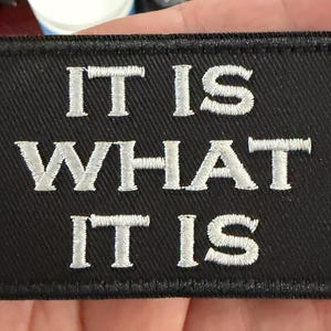 May include: Black rectangular patch with the embroidered phrase "IT IS WHAT IT IS" in white capital letters. The patch has a black border and is likely made of fabric.
