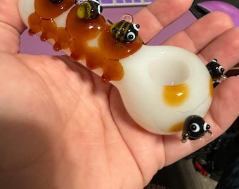 May include: A white glass pipe with a brown swirl design and four bee figurines attached to the side. The bees are made of glass and have black eyes and yellow and black stripes.