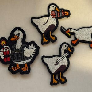 May include: Embroidered patches featuring cartoon geese in various action poses. One goose holds dynamite, another a knife, and a third carries a bundle of dynamite. The patches have white, brown, orange, and black details.