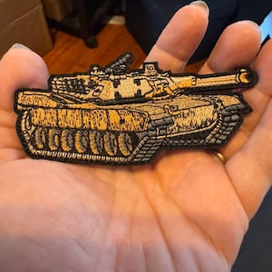 May include: Embroidered patch featuring a detailed depiction of a military tank. The patch showcases a tan and black color scheme, with intricate stitching that highlights the tank's design. The patch is held in a hand.