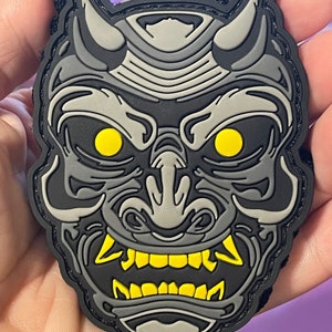 May include: A black and gray embroidered patch with a detailed design of a Japanese oni mask. The mask has yellow eyes and teeth, and a black background.