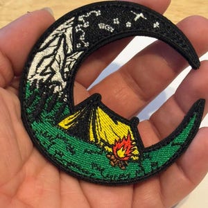 May include: Embroidered crescent moon patch featuring a camping scene. The patch has a black border, with a yellow tent, a campfire, and a starry night sky. The patch is green, black, white, yellow, and red.