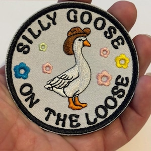May include: Embroidered patch featuring a white goose wearing a brown cowboy hat, with the text "SILLY GOOSE ON THE LOOSE" in a circular design. The patch includes colorful floral accents and a black border.