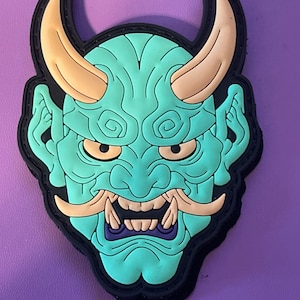May include: A green and orange Japanese demon mask patch with a fierce expression. The mask has sharp teeth, horns, and swirling patterns.