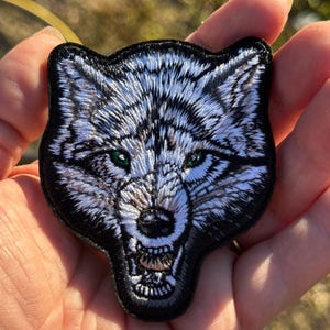 May include: Embroidered patch depicting a detailed wolf head with a snarling expression. The wolf has white and grey fur, green eyes, and a black border. Suitable for jackets, bags, or other fabric items.