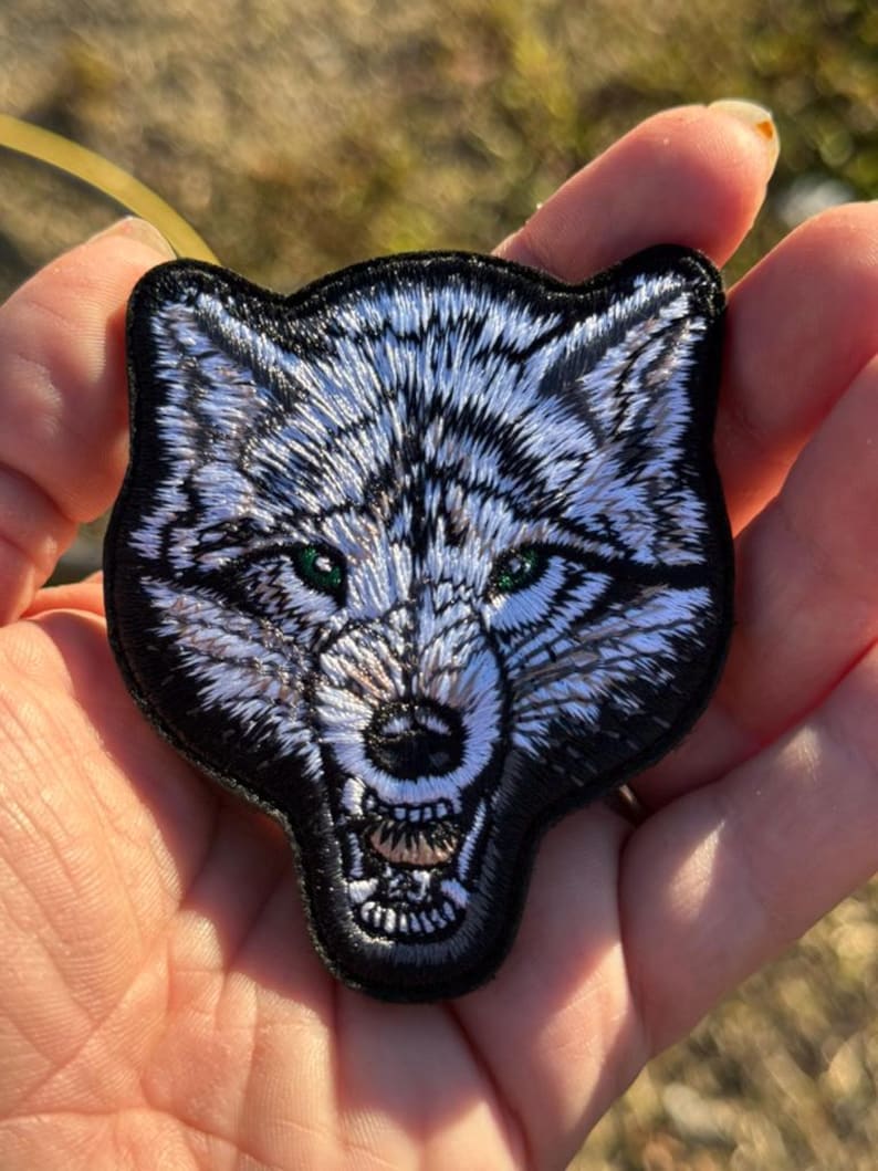 May include: Embroidered patch depicting a detailed wolf head with a snarling expression. The wolf has white and grey fur, green eyes, and a black border. Suitable for jackets, bags, or other fabric items.