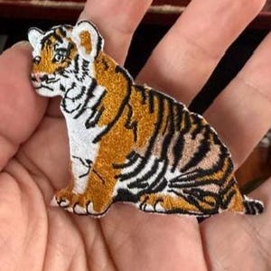 May include: Embroidered patch of a sitting tiger cub. The patch features a detailed design with orange, black, and white colors. The tiger cub is depicted with its paws forward, and its body is turned to the side.