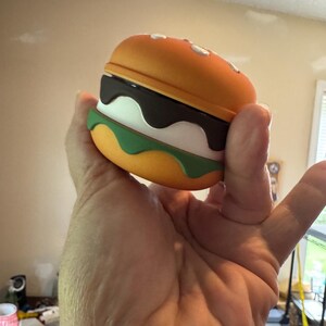 May include: A novelty burger-shaped case, featuring a bright orange top and bottom bun with white sesame seeds. The burger has layers of dark brown, white, and green, resembling a hamburger. The case is held in a hand.