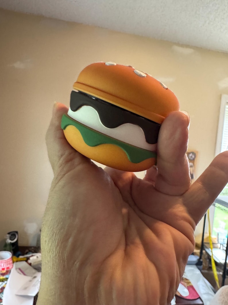 May include: A novelty burger-shaped case, featuring a bright orange top and bottom bun with white sesame seeds. The burger has layers of dark brown, white, and green, resembling a hamburger. The case is held in a hand.