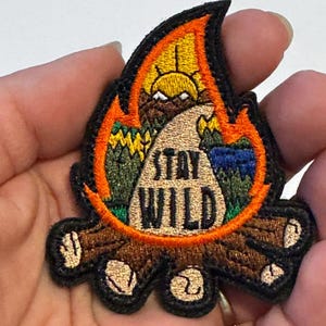 May include: Embroidered patch in the shape of a campfire with the words "Stay Wild". The patch features a sunrise, trees, and a road. The flames are orange and yellow, and the logs are brown.