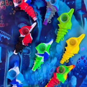 May include: A set of eight colorful silicone seahorse pipes. Each pipe features a different color combination and a clear glass bowl. The pipes are arranged on a blue and green background.