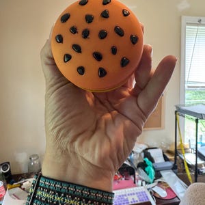 May include: A round, orange cookie-shaped stress ball with black chocolate chip details. The stress ball is held in a hand, with a turquoise and black patterned bracelet visible on the wrist.