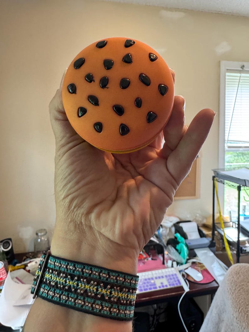 May include: A round, orange cookie-shaped stress ball with black chocolate chip details. The stress ball is held in a hand, with a turquoise and black patterned bracelet visible on the wrist.