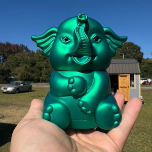 May include: A metallic emerald green elephant figurine with a smiling face, large ears, and a curled trunk. The figurine is held in a hand against a backdrop of a blue sky and green grass.