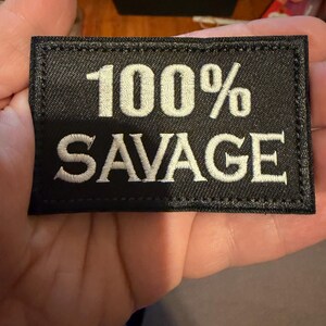 May include: Black rectangular patch with white embroidered text reading "100% SAVAGE." The patch has a stitched border and is held in a hand. The text is in a bold, sans-serif font.