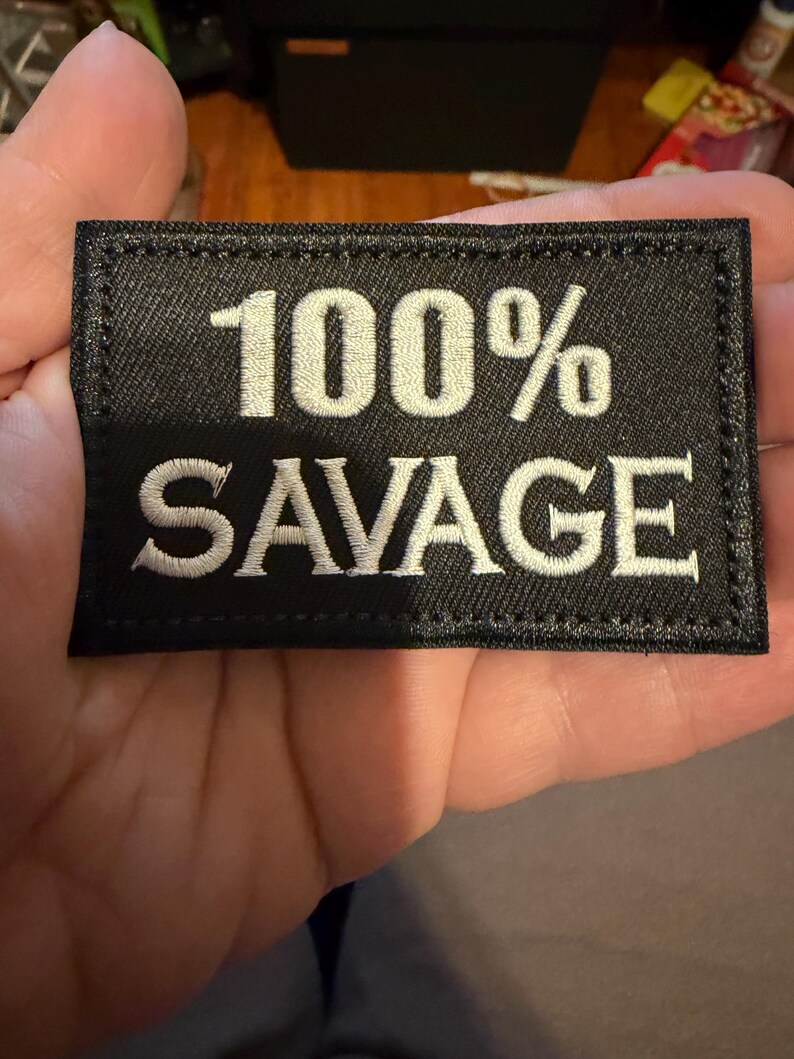 May include: Black rectangular patch with white embroidered text reading "100% SAVAGE." The patch has a stitched border and is held in a hand. The text is in a bold, sans-serif font.