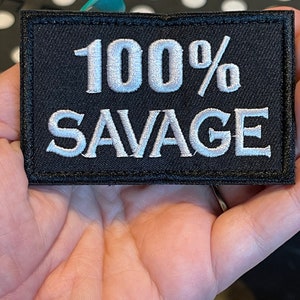 May include: Black rectangular patch with white embroidered text that reads "100% SAVAGE". The patch is held in a hand. The patch has a black border.