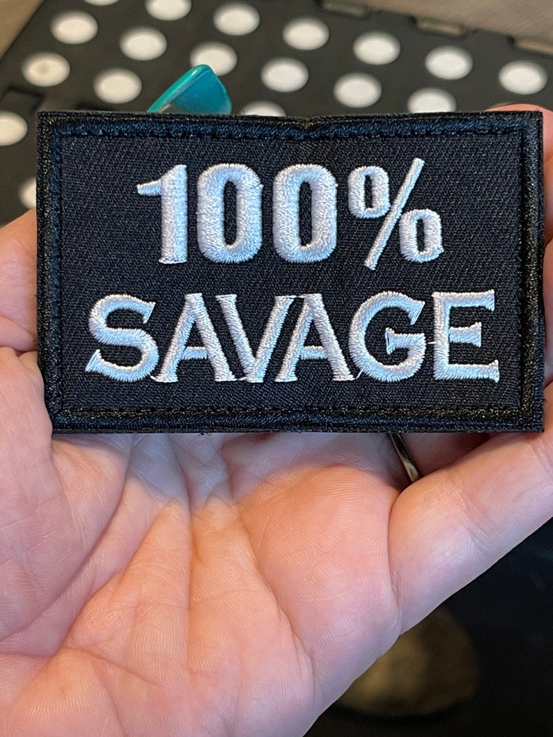 May include: Black rectangular patch with white embroidered text that reads "100% SAVAGE". The patch is held in a hand. The patch has a black border.