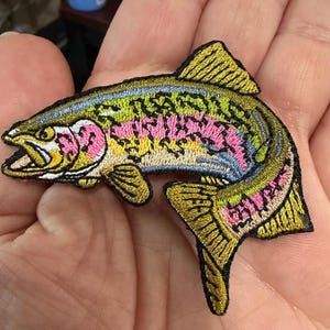 May include: Embroidered patch of a rainbow trout. The fish features a detailed design with a yellow, green, pink, and blue color scheme. The patch is held in a hand, showcasing the intricate stitching and vibrant colors.