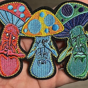 May include: Three embroidered mushroom patches with different colors and designs. The first mushroom is red and pink with blue and white details. The second mushroom is yellow and blue with orange details. The third mushroom is green and blue with purple details.